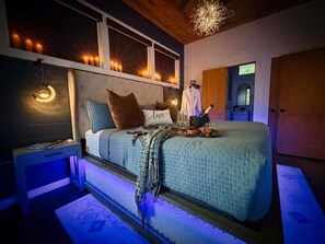 A bedroom with a bed, bedside tables, a nightstand, a decorative wall with candles, a chandelier, and a window.