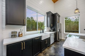 A modern kitchen with dark cabinetry, white countertops, and large windows offering views of the outdoors.