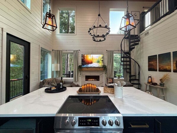 A modern kitchen with a central island, a built-in oven, and a stove. There is a fireplace with a TV above it, and a spiral staircase to the upper level.
