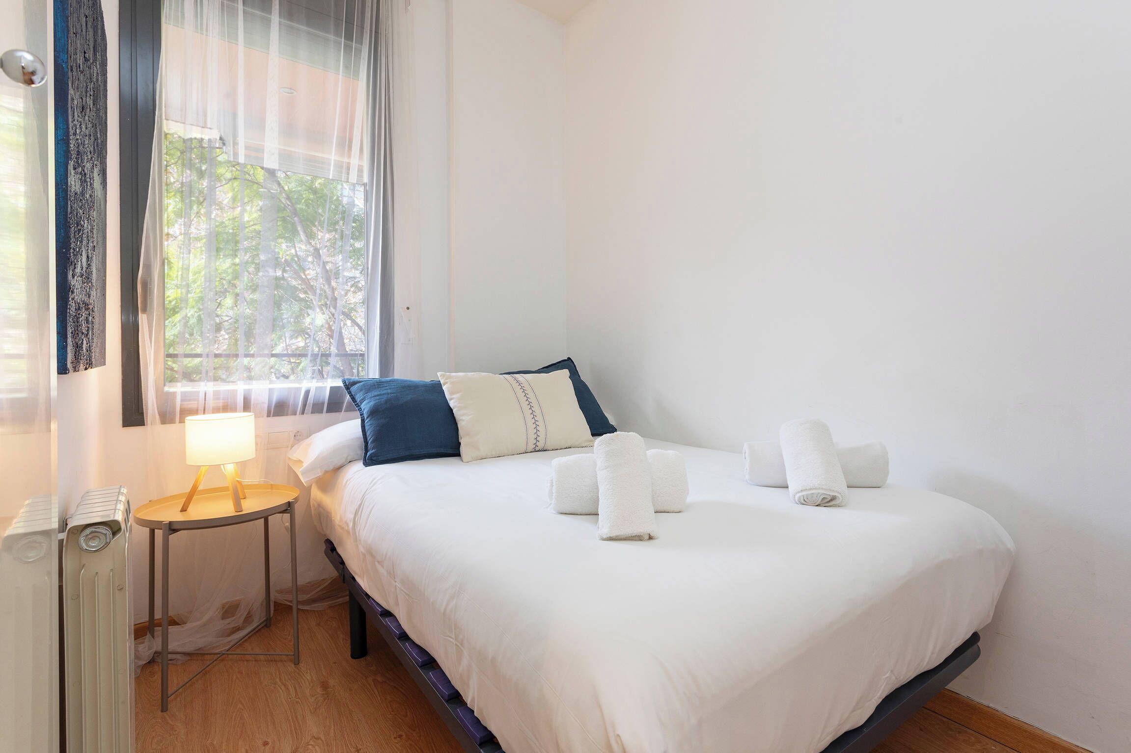 A neatly made bed with white linens and blue pillows, a bedside table with a lamp, and a window with sheer curtains.