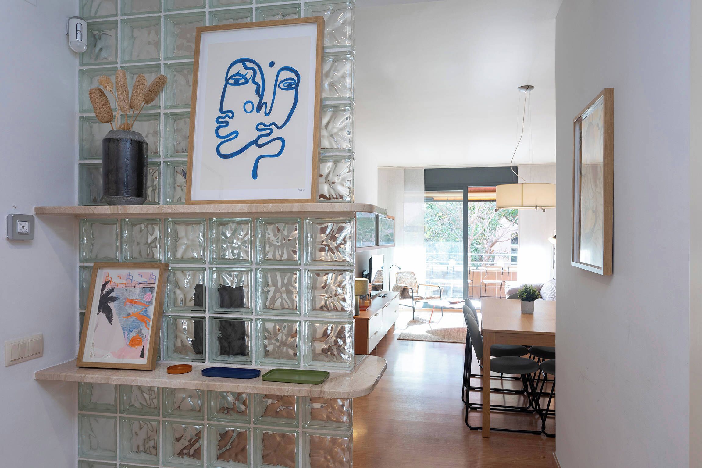 Een moderne keuken met een glazen scheidingswand, kunstwerken in lijsten, en een eethoek met een tafel en stoelen.