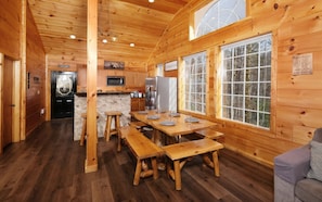 A cabin interior with wooden walls, a kitchen area, a dining table with benches, and large windows.