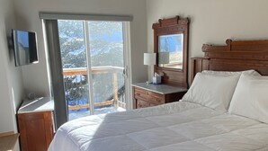 Ein Schlafzimmer mit einem großen Bett, einem Deckenventilator, einem Fernseher, einer Holzkommode, einem Nachttisch mit Lampe und einem Fenster mit Blick auf schneebedeckte Bäume.