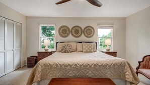 Quarto com cama, ventilador de teto, mesa de cabeceira, abajur, cadeira e janela com persianas.