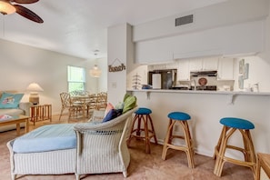 A living room with wicker furniture, a dining table, and a kitchen area with white cabinets and a countertop.