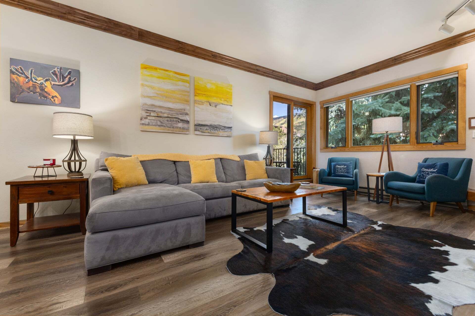 A living room with a gray sectional sofa, a wooden coffee table, and a cowhide rug. There are two armchairs and a lamp on a side table.