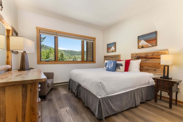 A bedroom with a bed, bedside table, lamp, and a window with a view of greenery.