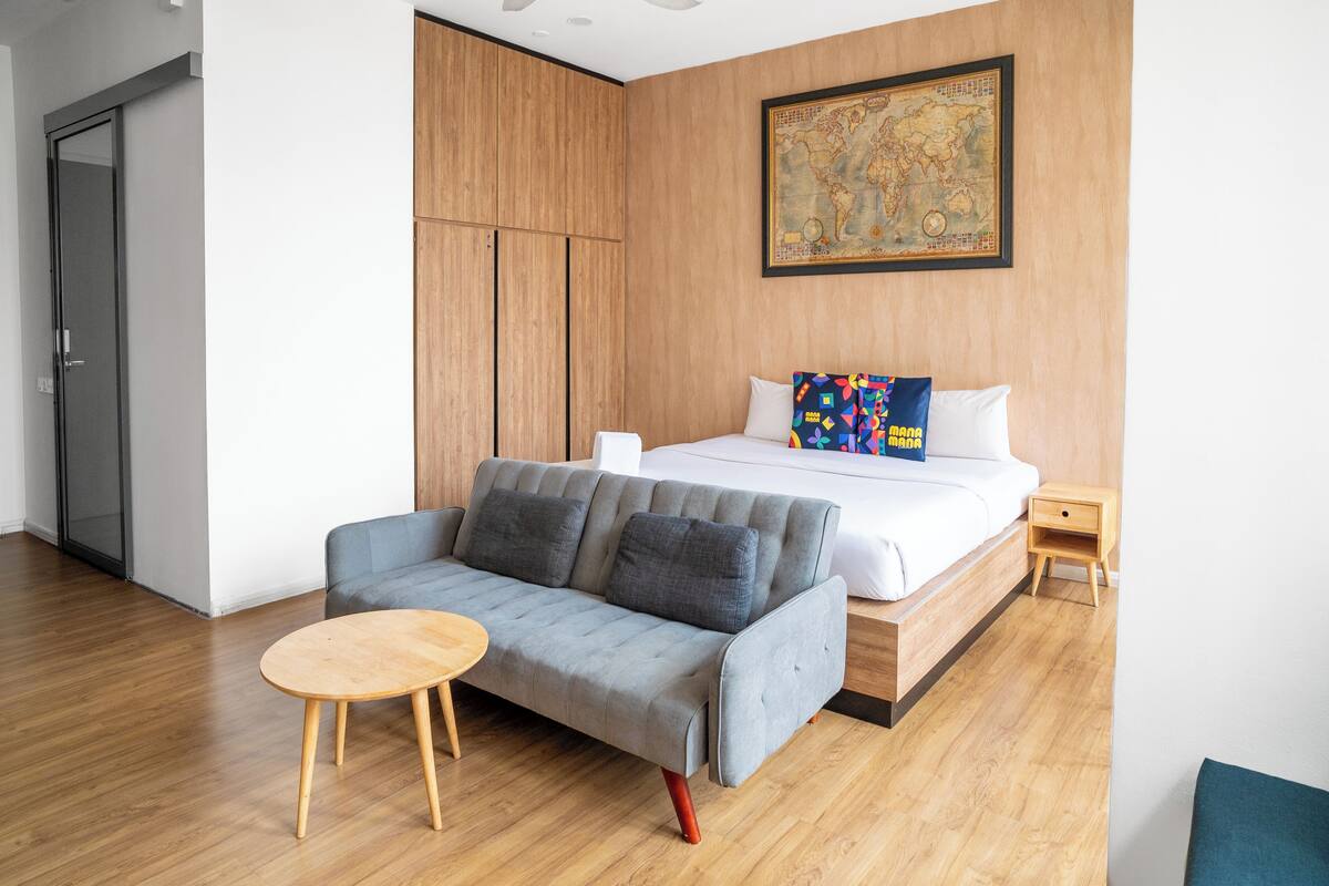 Una habitació d'hotel moderna amb un sofà gris, un terra de fusta, un llit amb roba de llit blanca, una petita taula de nit de fusta i un mapa a la paret.