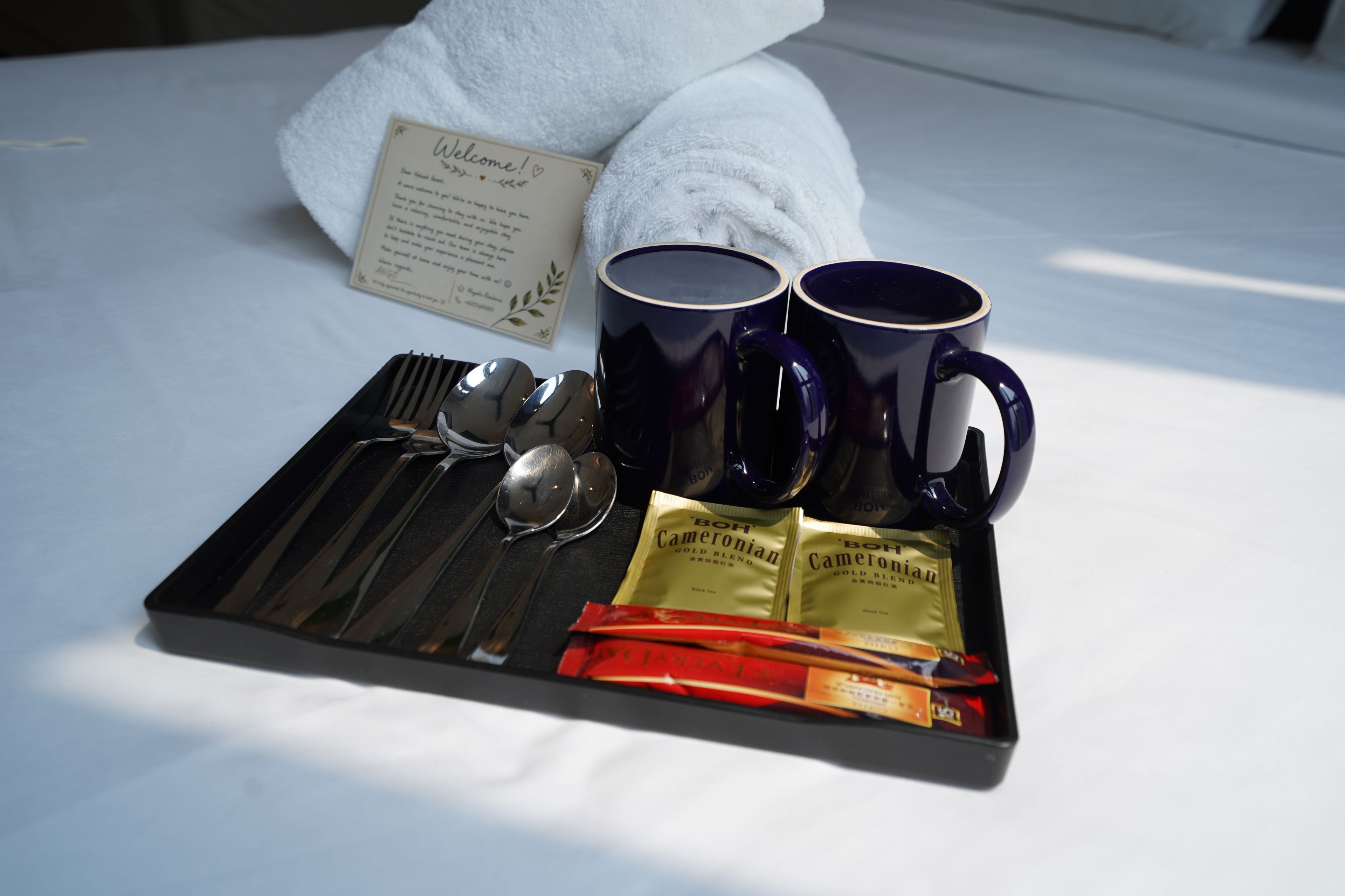 Een dienblad met twee donkere mokken, een lepel, een vork en twee zakjes 'BOH' Cameronian Gold Blend thee, op een bed met een witte handdoek en een welkomstkaart.