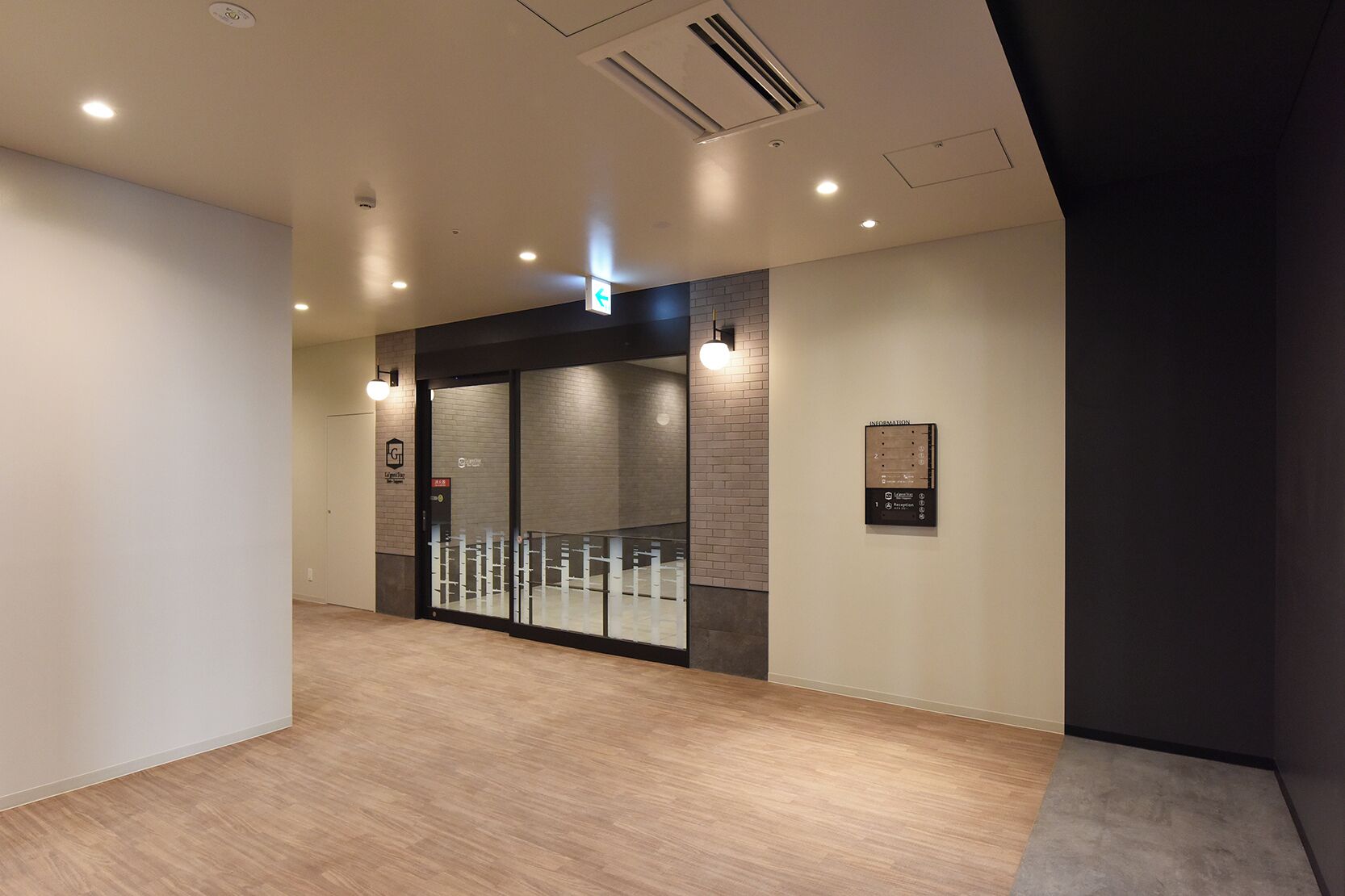 A modern hotel lobby with a wooden floor, a glass door entrance, and a wall-mounted information board.