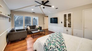 Ein geräumiges Wohnzimmer mit einem großen Bett, einem Sofa, einem Couchtisch und einem Deckenventilator. Es gibt Fenster mit Jalousien, einen an der Wand montierten Fernseher und eine Vitrine mit Glastüren.