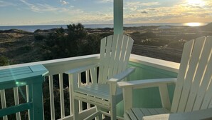 A covered outdoor seating area with two white Adirondack chairs and a small table, overlooking a beach and ocean view during sunset.