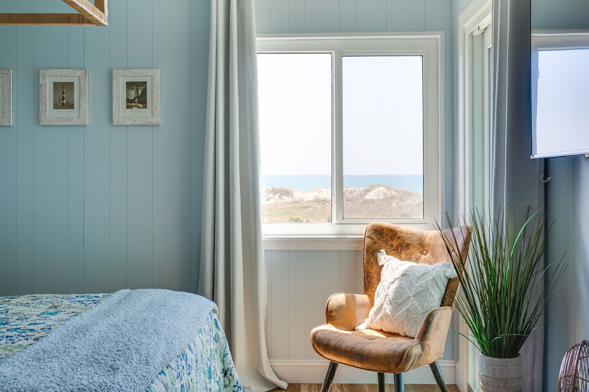 A bedroom with a large window, a bed with a patterned bedspread, a chair with a white pillow, and a potted plant.