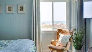 A bedroom with a large window, a bed with a patterned bedspread, a chair with a white pillow, and a potted plant.