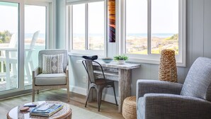 A room with a view of the ocean, a wooden desk, two chairs, and a round table with books.