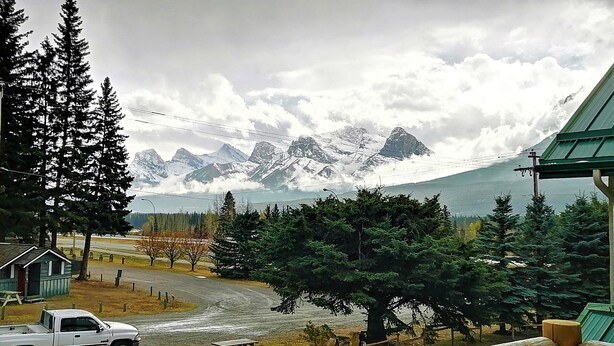 Vista de un paisaje montañoso con picos nevados, una cabaña de madera, una camioneta pickup blanca y una mesa de picnic.
