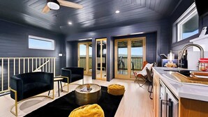 A modern living room with a black sofa, a coffee table, and a yellow ottoman. There is a kitchen area with a sink and a window. - Sleeps 20 , Game Barn, Pool, Hot Tub, Luxe Retreat (Dripping Springs)