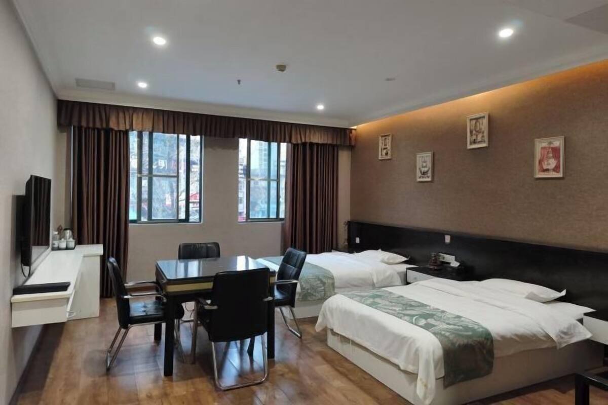 A hotel room with a desk, two beds, a TV, and large windows with curtains.