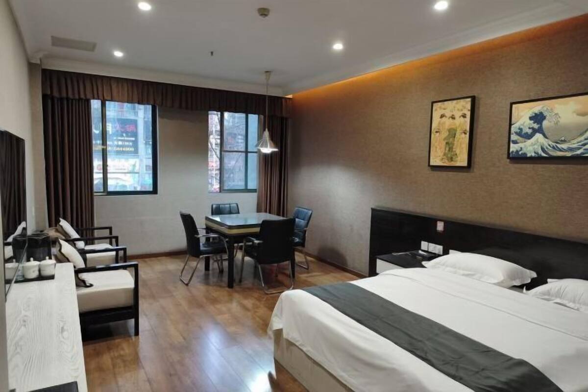 A hotel room with a bed, a desk, and a chair. There are two windows with curtains, and artwork on the wall.