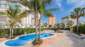 Zona de piscina con palmeras, sillas de descanso y un edificio al fondo.