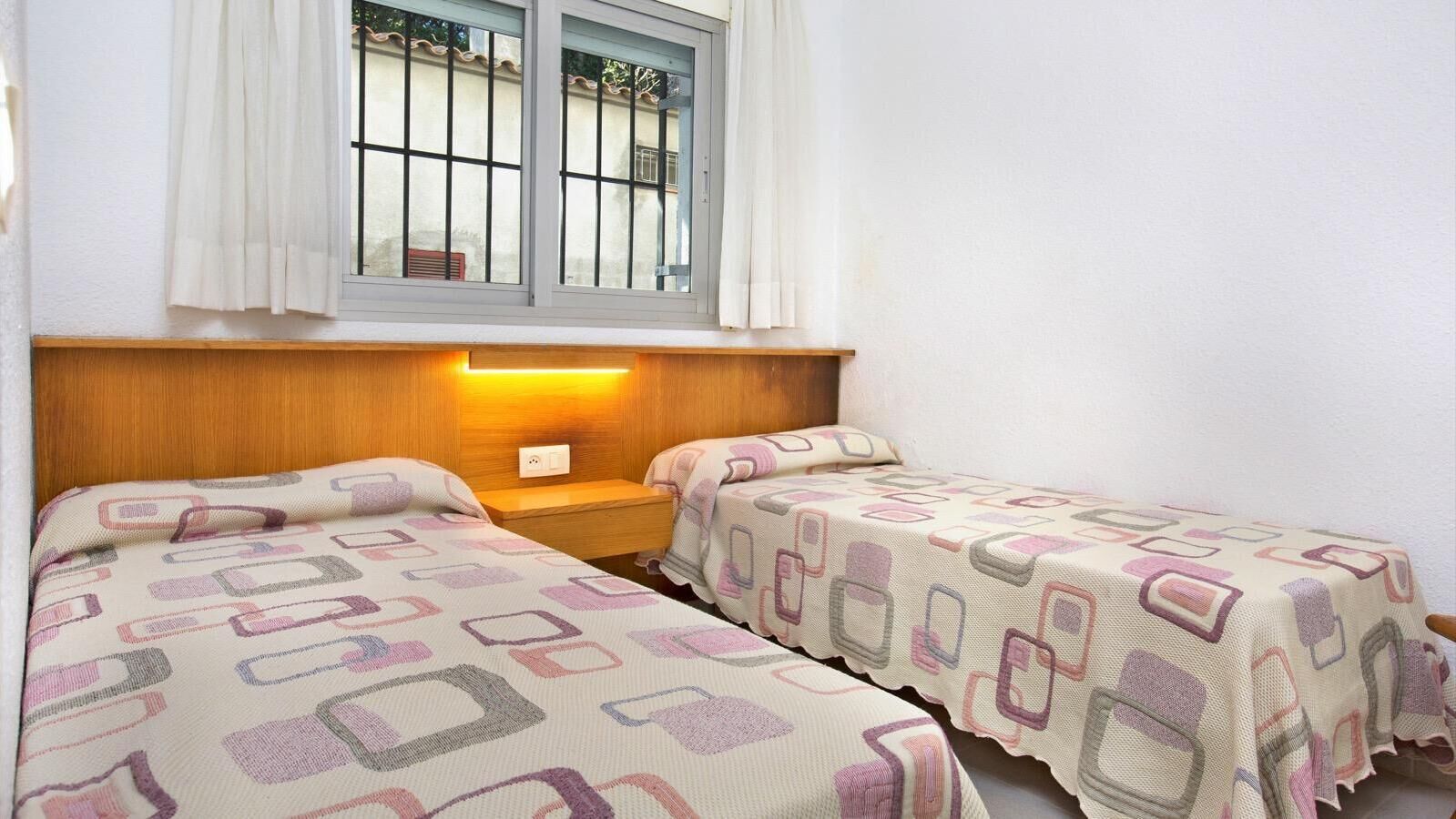 A room with two beds, a nightstand, a window with curtains, and a wall-mounted light.