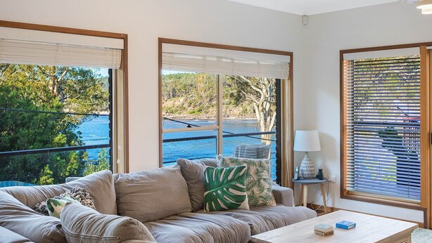 A living room with a sofa, a coffee table, and a view of a body of water and greenery. - Gillmith - Up to 10 people (Pambula Beach)