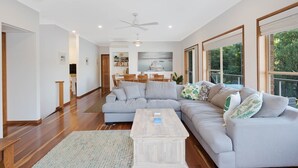 A spacious living room with a large grey sofa, a coffee table, and a dining area in the background. - Gillmith - Sleeping up to 16 people (Pambula Beach)