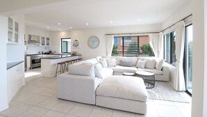 A modern living room with a large L-shaped sofa, a small round coffee table, and a kitchen area with white cabinets and a built-in oven. - Villa Paradiso - Pool and Beach Front (Tura Beach)