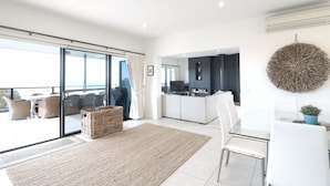 A modern interior with a glass dining table, white chairs, a kitchen area, and a balcony with wicker furniture. - Villa Paradiso - Pool and Beach Front (Tura Beach)