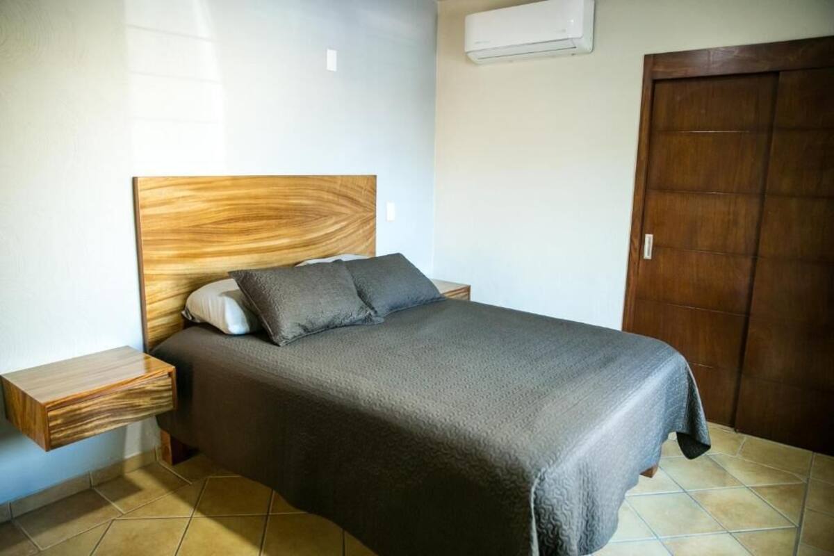 Un dormitorio con cabecera de madera, cama con manta gris, mesita de noche y armario de madera.