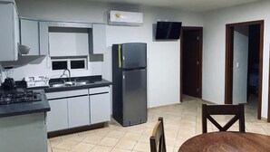 A compact kitchen with a gas stove, sink, and a refrigerator. There is a flat-screen TV mounted on the wall and a dining area with a wooden table and chairs.