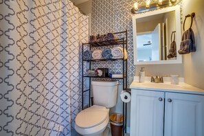 A bathroom with a patterned shower curtain, a toilet, a sink with a mirror, and a shelf with towels and toiletries. - Immaculate 2-Bed in Dallas Close to Everything! (Dallas)