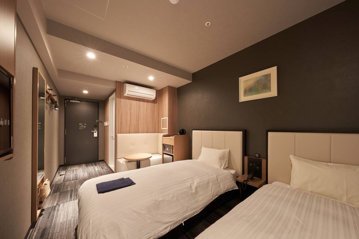 Habitación de hotel con dos camas, una mesita redonda pequeña, un escritorio con una silla y un equipo de aire acondicionado instalado en la pared.