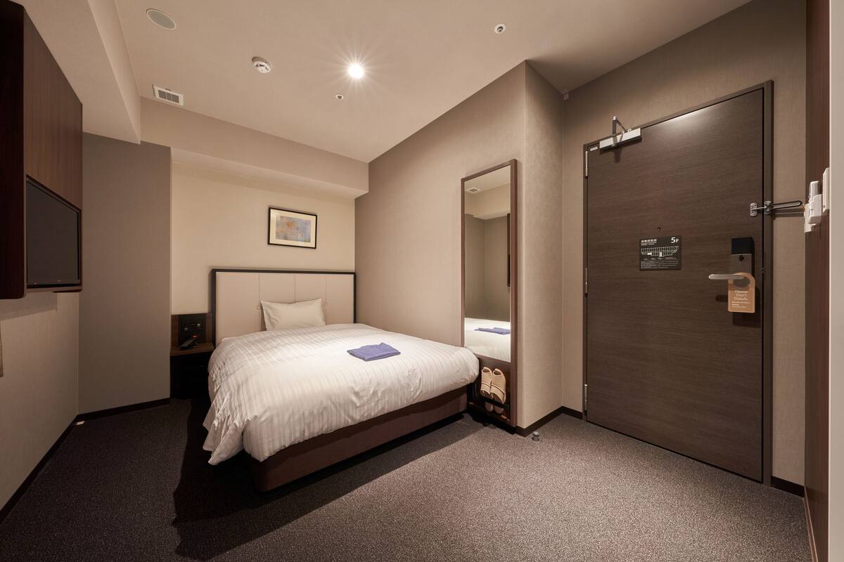 Una habitación de hotel con cama, mesita de noche, espejo y puerta con un cartel.