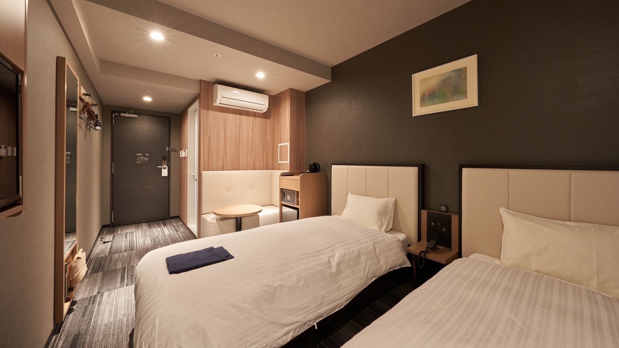 A hotel room with two beds, a small table, a chair, an air conditioning unit, and a painting on the wall.