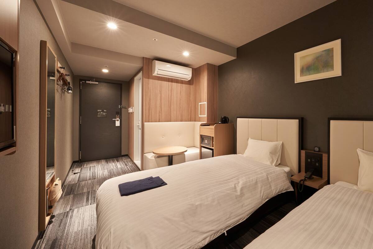 Habitación de hotel con dos camas, una mesita, una silla, un equipo de aire acondicionado y un cuadro en la pared.