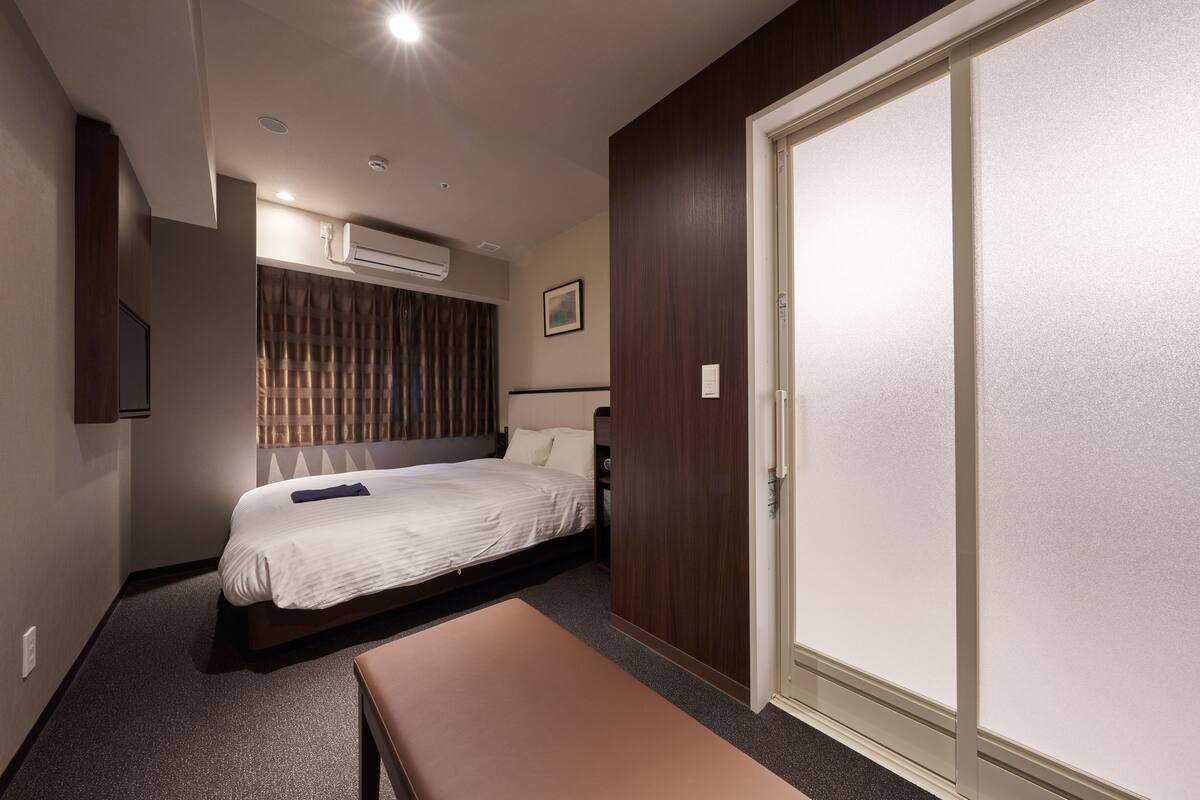 Una habitación de hotel con cama, una banca, un equipo de aire acondicionado, un armario y una puerta.