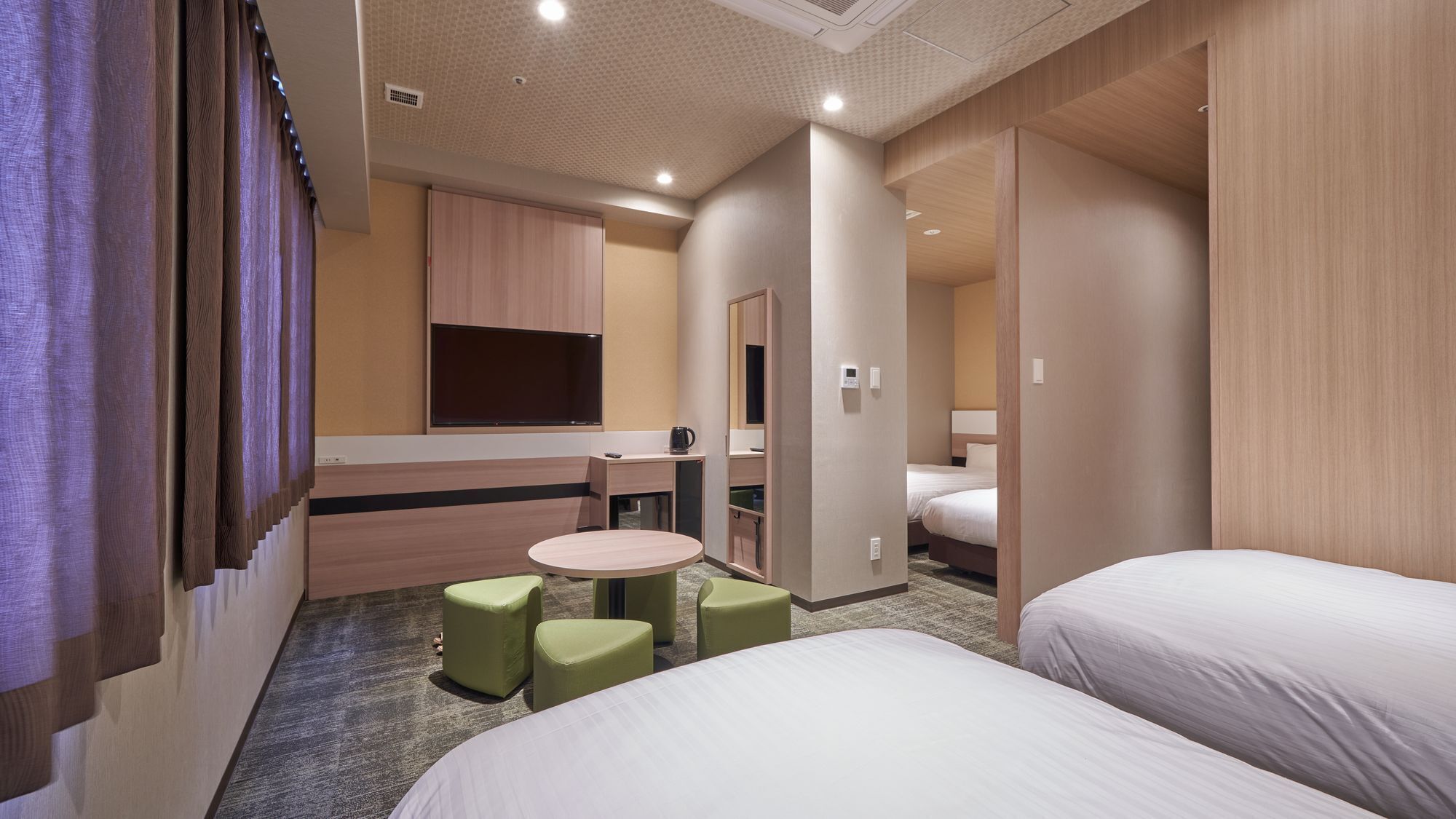 A hotel room with two beds, a small round table, two green chairs, a minibar, and a mirror.