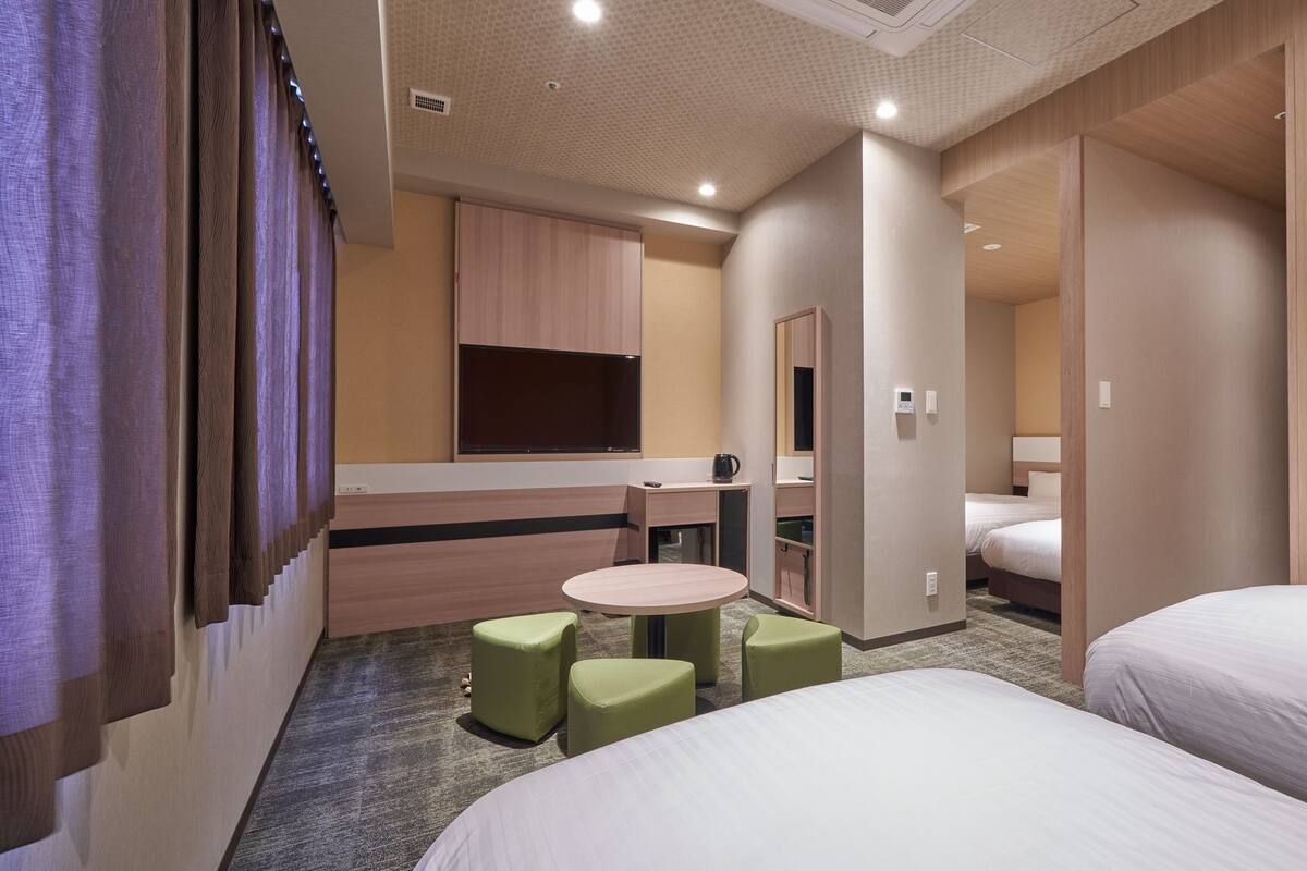 Habitación de hotel con dos camas, una mesita redonda pequeña, dos sillas verdes, un minibar y un espejo.