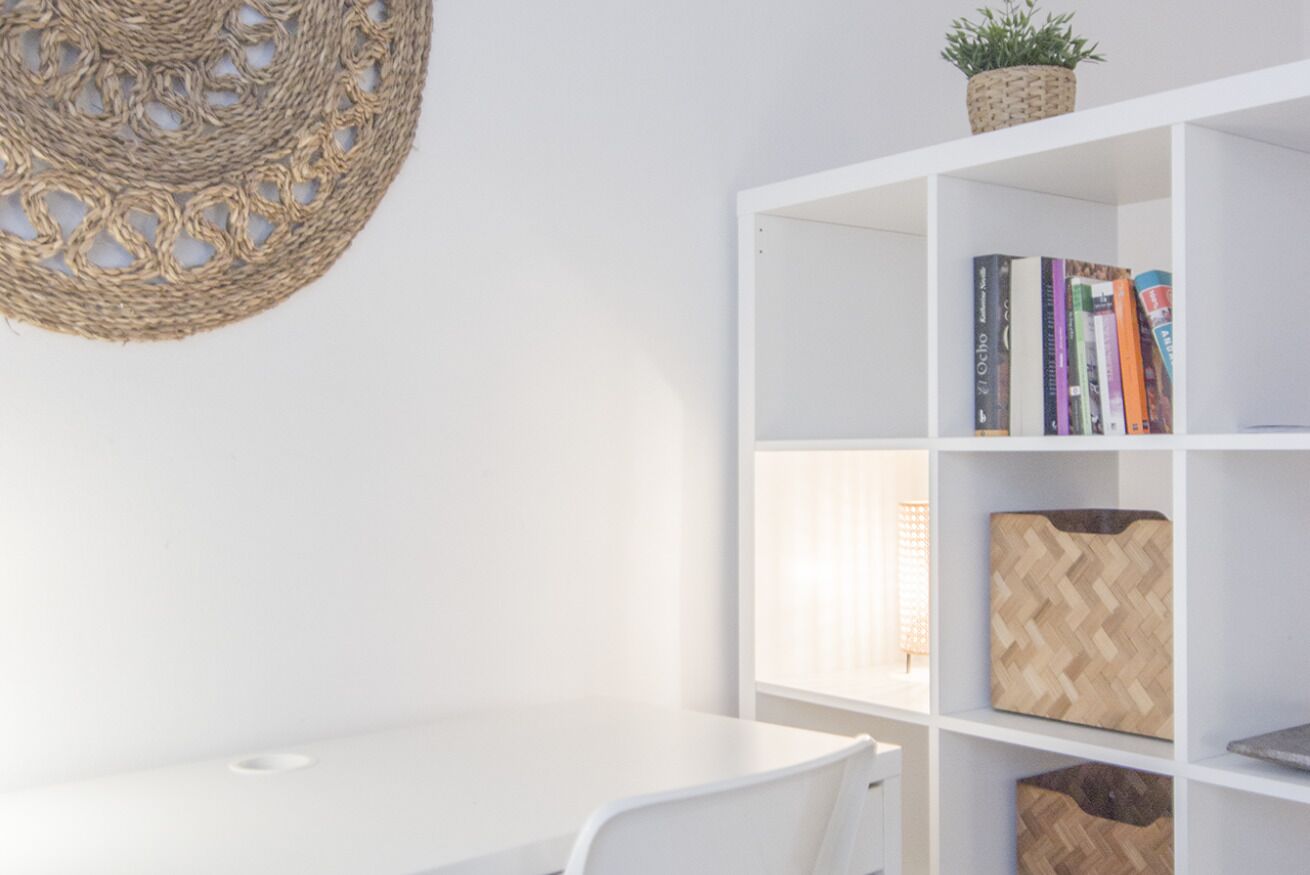 A white room with a woven wall hanging, a white bookshelf with books and decorative items, and a white desk.