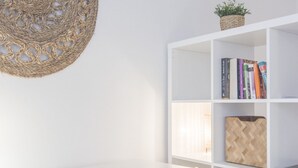 A white room with a woven wall hanging, a white bookshelf with books and decorative items, and a white desk. - Rentalsevilla Living La Alameda (Sevilla)