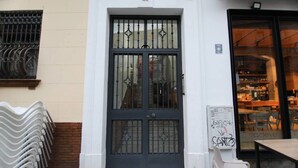 A white building with a black metal door and a window with metal bars. There is a white chair outside and a table with chairs inside. - Rentalsevilla Vive el Salvador (Sevilla)
