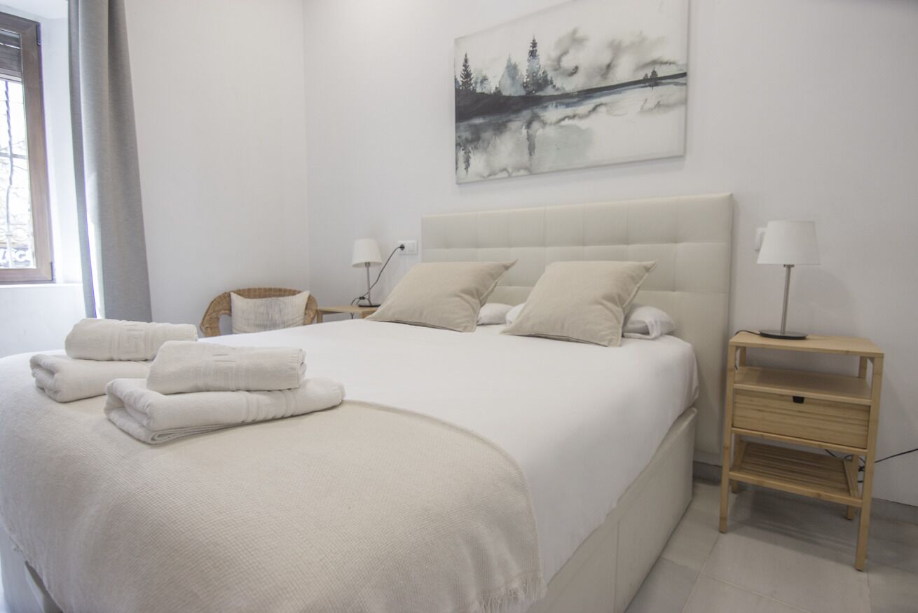 A neatly made bed with white linens and pillows, a bedside table with a lamp, and a painting of a forest on the wall.