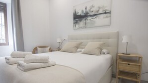 A neatly made bed with white linens and pillows, a bedside table with a lamp, and a painting of a forest on the wall. - Rentalsevilla Vive el Salvador (Sevilla)