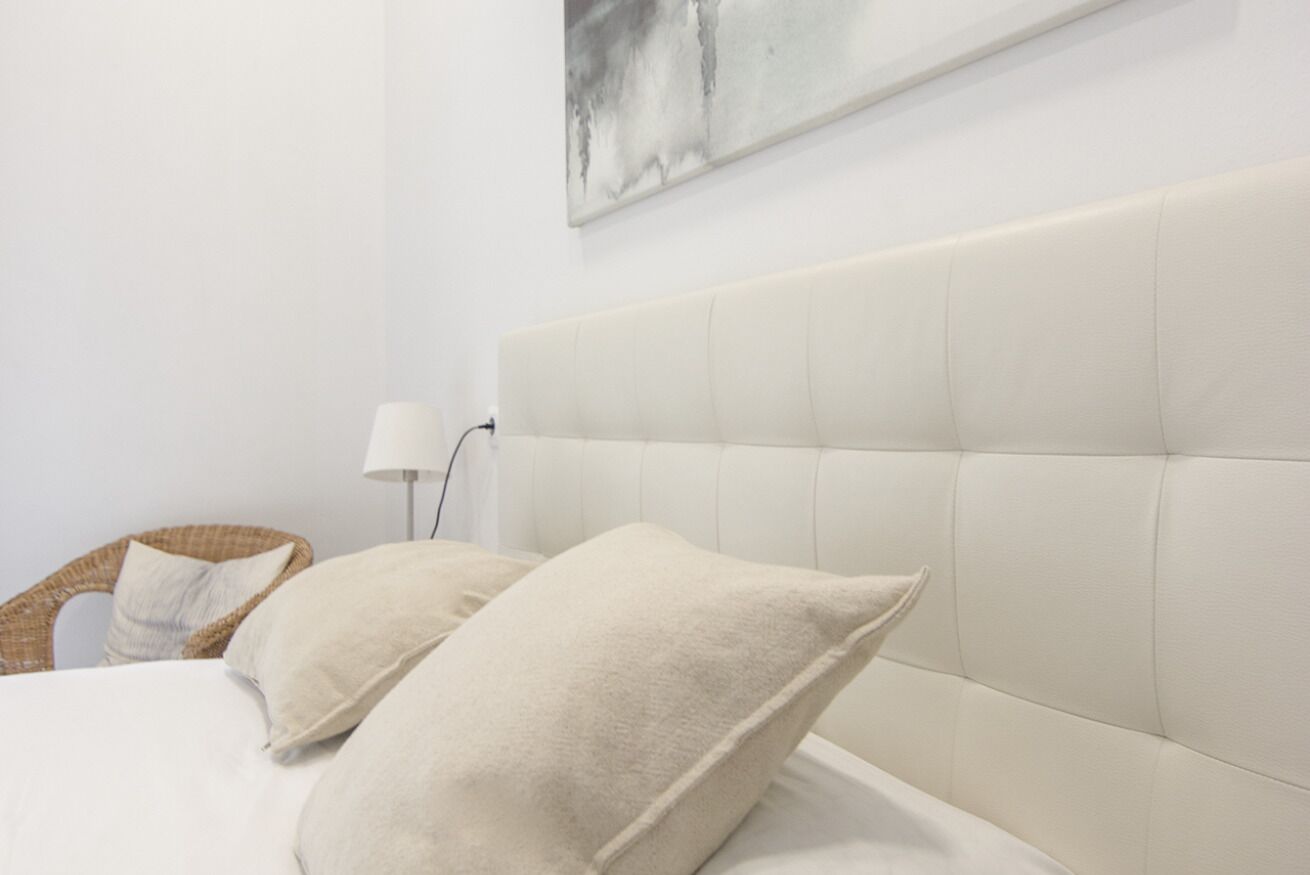 A neatly made bed with two pillows, a headboard, a wall-mounted lamp, and a wicker chair.