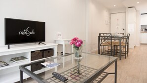 A modern living room with a flat-screen TV, a glass coffee table, a vase with flowers, a remote control, and a dining area with a table and chairs. - Rentalsevilla 3 Dormitorios Junto a la Macarena (Sevilla)