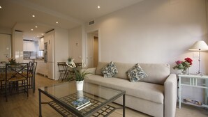 A modern living room with a glass coffee table, a beige sofa with patterned cushions, and a dining area with a wooden table and chairs. - Rentalsevilla 3 Dormitorios Junto a la Macarena (Sevilla)