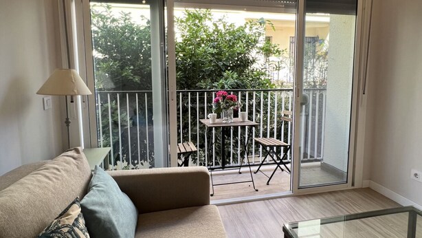 A balcony with a sliding glass door, a small table with flowers, and a sofa. - Rentalsevilla 3 Dormitorios Junto a la Macarena (Sevilla)
