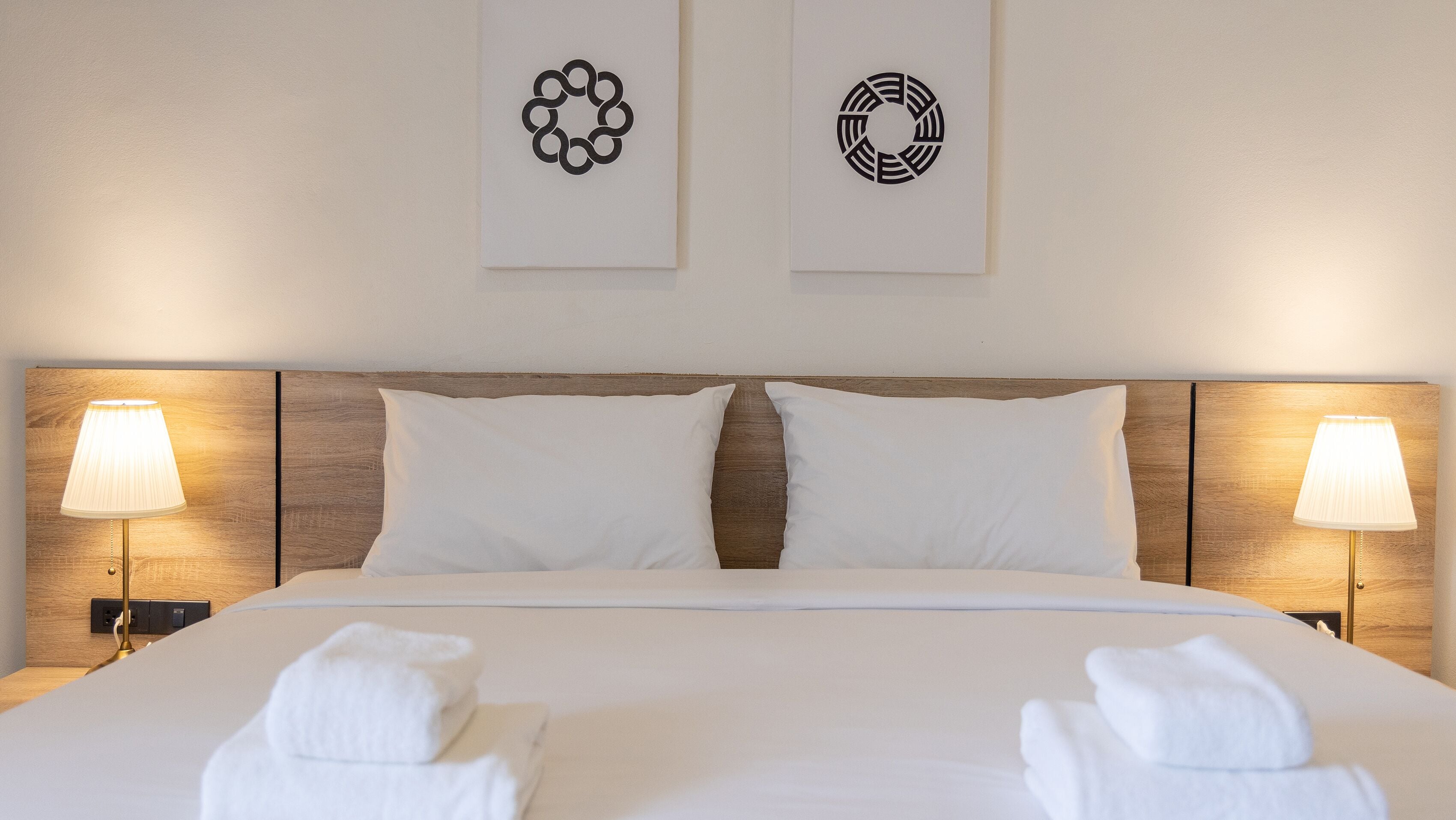 A neatly made bed with white linens and two pillows, flanked by two bedside lamps. Two towels are folded at the foot of the bed.