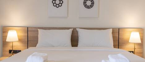 A neatly made bed with white linens and two pillows, flanked by two bedside lamps. Two towels are folded at the foot of the bed.